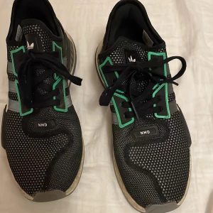 Adidas NMD sneakers svart/grön - Svarta Adidas NMD sneakers med grå och gröna detaljer samt vita och röda inslag. Ovandel i mesh med mönstrad struktur, svarta snören och klassiska tre ränder på sidan. Sulan är vit med svarta och grå partier, och baksidan har röd Adidas-logga.