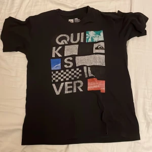 Svart Quiksilver t-shirt med tryck - Svart t-shirt från Quiksilver med stort grafiskt tryck framtill i flera färger och mönster, bland annat rutor och palmer. Klassisk passform med rund hals och korta ärmar. Materialet är en mjuk blandning av bomull och polyester.
