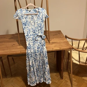 Blommig omlottklänning från Indiska - Långklänning från Indiska i vitt med blått blommönster. Omlottmodell med knyt i midjan, korta ärmar och v-ringad hals. Lätt och flowy känsla, perfekt för varma dagar. Materialet känns som bomull eller viskos.