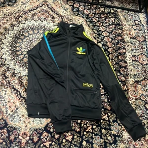 Adidas svart track jacket XS - Svart Adidas track jacket med färgglada ränder i gult och blått och grönt längs ärmarna. Jackan har dragkedja framtill, hög krage och broderad Adidas-logga i gult och blått o grönt på bröstet. CHILE 20-detalj vid fickan och klassisk sportig look.