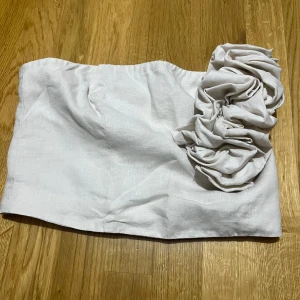 Vit tub med volang - Superfin vit bandeau tubtopp med stor volangdetalj på ena sidan. Toppen är axelbandslös och har en stilren, modern look som passar perfekt till sommarens outfits. Gjord i ett lätt och luftigt tyg som känns skönt mot huden.