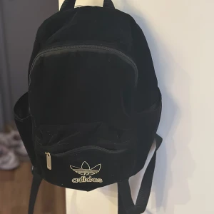 Svart Adidas ryggsäck med guldlogga - Snygg svart ryggsäck från Adidas med ikonisk guldlogga framtill. Väskan har flera fack med dragkedja, justerbara axelband och mesh på baksidan för extra komfort. Tillverkad i slitstark polyester, perfekt för skola eller cityhäng.