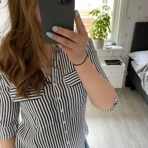 Randig vit och blå skjorta med knappar - Snygg skjorta med vita och mörkblå vertikala ränder, klassisk krage och knäppning framtill. Skjortan har långa ärmar som kan vikas upp och fästas med knapp. Bröstficka på båda sidor och lätt figursydd passform. Perfekt till jeans för en snygg look. Sparsamt använd och fint skick. Storlek XS