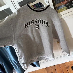 Grå sweatshirt med Missouri-tryck - Grå sweatshirt med svart och vitt Missouri-tryck på bröstet. Tröjan har rund halsringning med svart kant och långa ärmar. Den har ett litet skönhetsfel därav priset men det är en lite mörkare fläck vid höften som knappt syns.