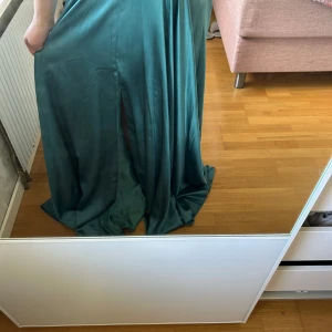 Mörkgrön långklänning med paljetter och snörning - Säljer en mörkgrön  långklänning med glittriga paljetter och broderier upptill. Klänningen har smala axelband, öppen rygg med snörning och en slits framtill. Perfekt för bal eller fest! Kjolen är i ett mjukt, glansigt material som faller snyggt. Den är endast använd en gång och är helt som ny. Nypris ligger på 6000kr