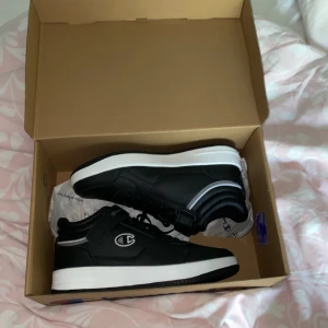 Svarta sneakers från Champion - Säljer ett par helt oanvända svarta sneakers från Champion med vita sulor och svarta snören. Skorna har Champion-loggan på sidan och bak, samt en klassisk rund tå och platt sula. Perfekta för en sportig och stilren look.
