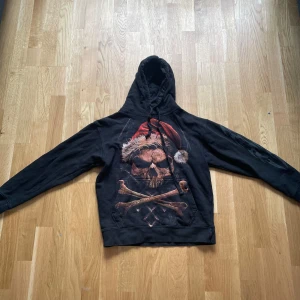 Svart hoodie med dödskalle och tomteluva - Säljer en svart hoodie med tryck av en dödskalle med tomteluva och korslagda benknotor på magen. Ärmarna har gråa flam-mönster. Perfekt för dig som gillar lite tuffare stil och vill sticka ut.