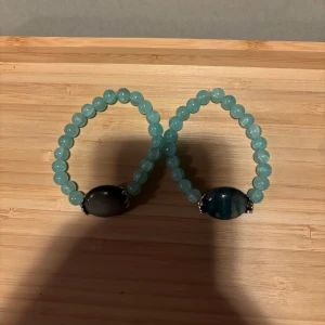 Blågröna pärlarmband (25kr/st) - (25kr/st) Två elastiska armband med blågröna runda pärlor och en större svart oval sten i mitten. Perfekt accessoar för att ge en färgklick till din outfit.