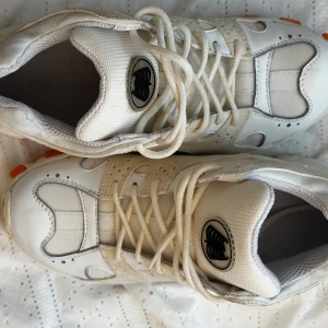 Vita sneakers från Our Legacy med orange Vibram-sula - Säljer ett par vita sneakers från Our Legacy med robusta orange detaljer på Vibram-sulan. Skorna har snörning, mesh och läderpaneler samt en unik logga på plösen. Perfekta för dig som gillar stilrena men ändå sportiga skor.
