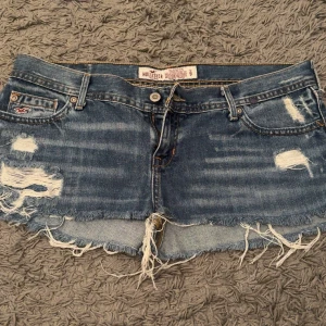 Blå slitna jeansshorts från Hollister - Säljer ett par blå jeansshorts från Hollister med slitna detaljer och fransig kant. Klassisk femficksmodell med låg midja och råa kanter. Perfekta för sommaren och en avslappnad stil. Storlek 9 skulle säga passar M, jag har S och de är lite stora