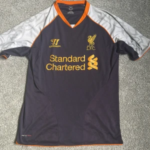 Liverpool FC tredjetröja 2013/14  - Säljer en Liverpool FC tredjetröja från säsongen 2013/14, tillverkad av Warrior. Tröjan är mörkgrå med vita och orange detaljer, korta ärmar och tryckt klubbmärke samt sponsorlogga på bröstet. Unikt mönster på ärmarna. Tröjan är i annvänt skick (se bild 5) men fortfarande bra skick. Den är äkta och köpt på classsic football shirts. Köparen står för frakt. Meddela mig om du har några frågor!