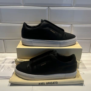 Arigato laceless - Dice Laceless Sneaker från Axel Arigato  Svart | Vit | Kalvläder | Slip-on modell | Guldfärgad logga | Räfflade sulor | Mått och passform: Normal passform | Material och skötsel: Yttre: Kalvläder 100% | Foder: Kalvläder 91% | NYPRIS 3000kr