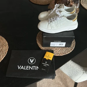 Vita sneakers från Valentino  - Säljer ett par snygga vita sneakers från Valentino med guldfärgad detalj på hälen. Skorna har klassisk snörning och rund tå. Kommer med originalkartong, dustbag och äkthetskort. Perfekta för dig som vill ha en stilren och lyxig look.
