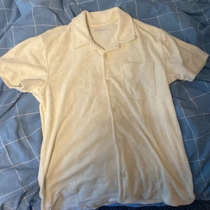 Beige frottéskjorta från Kappahl  - Säljer en beige kortärmad skjorta från Kappahl i mjuk frotté. Skjortan har klassisk krage, knäppning framtill och en bröstficka. Perfekt för en avslappnad och stilren look. Den passar även på folk som har M. Inget fel på tröjan bra skick.