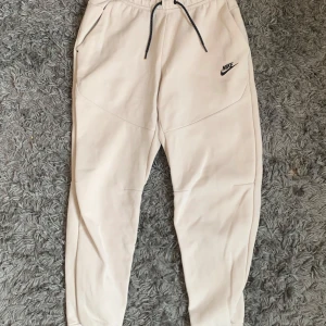 Beige mjukisbyxor från Nike med snörning - Snygga beige mjukisbyxor från Nike med svarta detaljer och snörning i midjan. Byxorna har fickor fram och en dragkedjeficka bak. Perfekta för en avslappnad stil eller till träning.