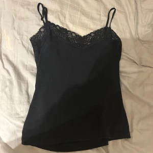 Svart linnetopp med spets - Svart linnetopp med tunna axelband och vacker spetsdetalj längs urringningen. Perfekt att styla till olika outfits för en trendig look.