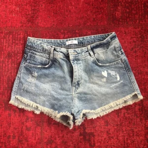 Blå jeansshorts med fransar och tryck - Säljer ett par blå jeansshorts med fransig kant och vita tryckdetaljer som hjärtan och texten 'HEART JOY'. Shortsen har klassisk femficksdesign och normal passform. Perfekta för sommardagar!