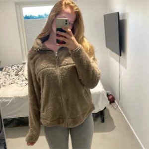 Beige/brun teddyjacka med dragkedja - Mysig beige teddyjacka med dragkedja framtill och hög krage. Jackan har lång ärm och en avslappnad passform, perfekt för kyligare dagar. Enkel och stilren design som passar till många outfits.