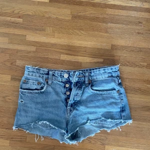 Blå jeansshorts med fransar från Zara - Säljer ett par blå jeansshorts från Zara med mid waist och råa fransade kanter. Shortsen har knappar framtill, klassiska fickor och ganska tajta 