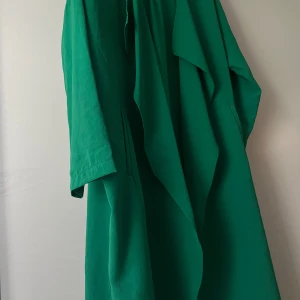 Grön omlottklänning från OsVet - Säljer en snygg grön omlottklänning från OsVet. Klänningen har trekvartsärm och en lös passform med fina draperade detaljer framtill. Perfekt för dig som vill ha en färgstark och bekväm look.