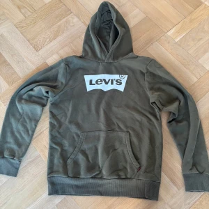 Olivgrön hoodie från Levi's - Säljer en olivgrön hoodie från Levi's med stor vit logga på bröstet. Tröjan har huva och en stor magficka framtill. Muddar vid ärmslut och nederkant. Perfekt för en avslappnad stil.