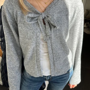 Grå kofta med rosettdetalj - Mysig grå kofta i mjukt material med lång ärm och stor rosett framtill. Perfekt att slänga över en topp för en avslappnad men ändå söt look. Enkel att matcha med jeans eller kjol.