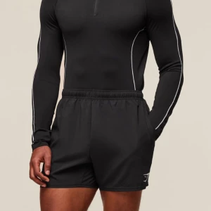 Svart långärmad träningströja från Gymshark - Säljer en svart långärmad träningströja från Gymshark med vit piping längs ärmar och sidor. Tröjan har en halv dragkedja framtill och är tillverkad i ett stretchigt syntetmaterial som passar perfekt för träning.