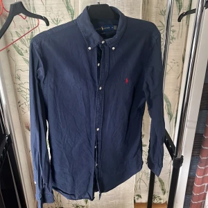 Mörkblå skjorta från Ralph Lauren - Säljer en klassisk mörkblå skjorta från Ralph Lauren i slim fit-modell. Skjortan har långa ärmar, knappar framtill och en broderad logga i rött på bröstet. Perfekt för dig som gillar stilrena plagg med snygga detaljer.