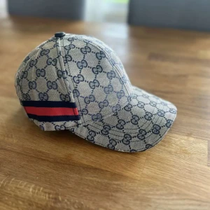 Beige keps från Gucci med GG-mönster - Snygg beige keps från Gucci med klassiskt GG-mönster över hela kepsen och en röd-blå rand på sidan. Kepsen har böjd skärm och justerbar passform. Perfekt för dig som vill ha en exklusiv accessoar.