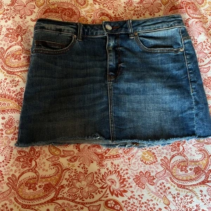 Jeanskjol med spetsdetalj och hjärta från Vero Moda - Säljer en blå jeanskjol från Vero Moda i storlek S. Kjolen har rå kant nertill, spetsdetalj på ena bakfickan och ett vitt hjärta på den andra. Klassisk femficksmodell med dragkedja och knapp framtill.