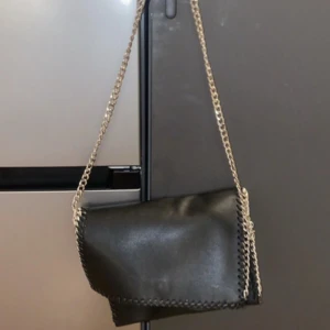 Svart axelväska med kedja från NLY Accessories - Säljer en svart axelväska från NLY Accessories med silverfärgad kedja som axelrem. Väskan har dekorativa sömmar längs kanterna och är tillverkad i syntetmaterial. Perfekt för att lyfta din outfit.