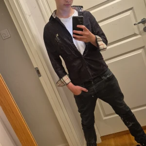 Svart Burberry skjorta  - Säljer en svart skjorta från Burberry med klassiska rutiga detaljer på insidan av kragen och ärmsluten. Skjortan har broderad logga på bröstet och knäppning framtill. Perfekt för dig som gillar stilrena och exklusiva plagg.