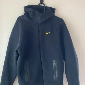 Nocta tech fleece - Svart hoodie med dragkedja från Nike NOCTA-kollektionen. Jackan har hög krage, två fickor med dragkedja och gul Nike-logga på bröstet. Snygga detaljer i gult även på ryggen. Perfekt för en sportig och stilren look.