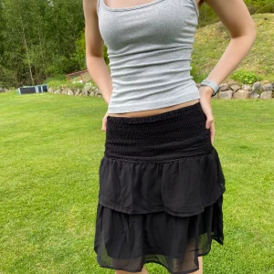 Svart volangkjol från ADPT - Kjolen är helt ny och oanvänd! Supersöt svart kortkjol från ADPT med smock i midjan och dubbla volanger. Kjolen är luftig och har en flowy känsla, perfekt till varma dagar. Tillverkad i ett tunt, lätt material som ger en snygg siluett. Går att vika ner för en kortare modell.