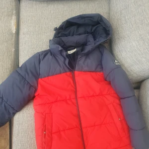 Röd och mörkblå pufferjacka med huva - Säljer en pufferjacka i rött och mörkblått med huva och dragkedja framtill. Jackan har två fickor på sidorna och quiltad design. Perfekt för kyliga dagar.