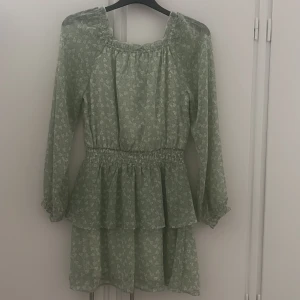 Grön blommig volangklänning - Söt ljusgrön klänning med små vita blommor och volangdetaljer. Klänningen har lång ärm, resår i midjan och dubbla volanger i kjolen. Perfekt för vår och sommar, med en luftig och lätt känsla.