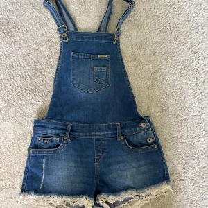 Jeansshorts med hängslen från Superdry - Säljer ett par blå jeansshorts med hängslen från Superdry. Shortsen har spetsdetaljer längst ner, klassiska fickor fram och bak samt en bröstficka. 