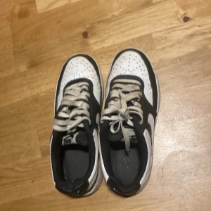 Nike Air Force 1 i svart och vitt - Säljer ett par klassiska Nike Air Force 1 sneakers i svart och vitt. Skorna har vit tå med perforeringar, svart ovandel och vit sula. Ikonisk Nike Swoosh på sidan och snörning framtill. Perfekta för en stilren vardagslook.