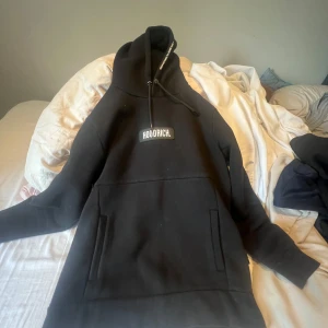 Svart hoodie från Hoodrich - Svart hoodie från Hoodrich med vit logga på bröstet och stor känguruficka framtill. Tröjan har huva med dragsko och långa ärmar. Perfekt skick, knappast använd för ett bra pris