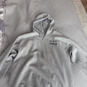 Grå hoodie från Gina Tricot med tryck - Grå hoodie från Gina Tricot med svart text 'JE M'EN AMOUR' och en stjärna tryckt på ryggen. Mjuk insida och klassisk huva. Perfekt för en avslappnad stil.