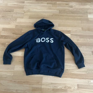 Mörkblå hoodie från BOSS - Säljer en mörkblå hoodie från BOSS med stor vit logga på bröstet. Tröjan har huva med dragsko och en stor magficka framtill. Perfekt för en avslappnad och stilren look.