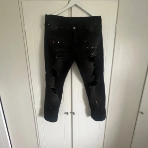 Svarta slitna jeans med färgstänk - Säljer ett par svarta jeans med stora slitningar på benen och coola färgstänk i orange och vitt. Jeansen har fem fickor och gylf. Perfekta för dig som gillar en edgy och unik stil. (Jeansen är custom alltså har jag gjort slitningarna och färgstänket själv, jeansen är väldigt lika ”purple jeans” man ser knappt nån skillnad.
