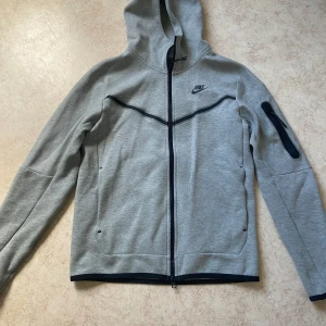 Grå hoodie från Nike - Säljer en grå hoodie från Nike med hel dragkedja och svarta detaljer. Hoodien har huva, lång ärm och två fickor framtill samt en extra ficka på ena ärmen. Perfekt för träning eller chill.