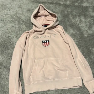 Ljusrosa hoodie från Gant - Säljer en ljusrosa hoodie från Gant med broderad logga och sköld på bröstet. Tröjan har huva och en stor magficka framtill. Perfekt för dig som gillar klassisk och sportig stil.