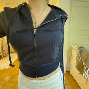 Svart croppad kofta med dragkedja - Säljer en svart croppad hoodie med dragkedja framtill och två stora fickor. Jackan har huva med dragsko och ribbade muddar i midjan. Perfekt för en avslappnad och trendig stil. 