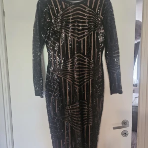 Svart glittrig långklänning från Boohoo - Säljer en svart långklänning från Boohoo med glittriga paljetter och geometriskt mönster. Klänningen har lång ärm och djup v-ringning i ryggen, perfekt för att sticka ut på festen.