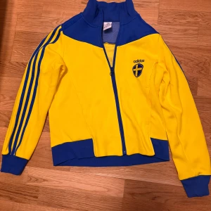 Gul och blå träningsjacka från Adidas - Säljer en gul och blå träningsjacka från Adidas med Sveriges landslagssköld på bröstet och texten 'Sweden' på ryggen. Jackan har dragkedja, lång ärm och klassiska Adidas-ränder på ärmarna.