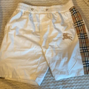 Vita shorts från Burberry med rutiga detaljer - Snygga vita shorts från Burberry med klassiskt rutigt mönster längs sidorna och broderad logga framtill. Elastisk midja med knappar och fickor både fram och bak. Perfekta för sommaren med en stilren och exklusiv look.