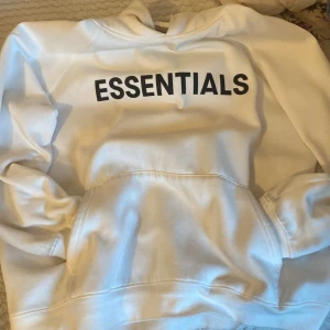 Vit hoodie från Essentials Fear of God - Säljer en vit hoodie från Essentials Fear of God med svart tryck på bröstet och ryggen. Tröjan har huva och en stor magficka framtill. Perfekt för en avslappnad och stilren look.