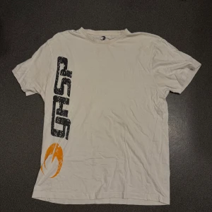 Vit t-shirt från GASP med tryck - Vit t-shirt från GASP med stort svart och orange tryck på framsidan och stor GASP-logga på ryggen. Klassisk passform och rund halsringning. Perfekt för dig som gillar sportig stil.
